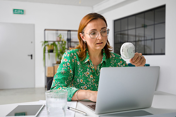 A woman holding a small fan while working on a laptop