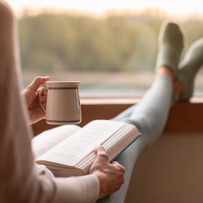 Person holding a mug and reading a book with feet propped up